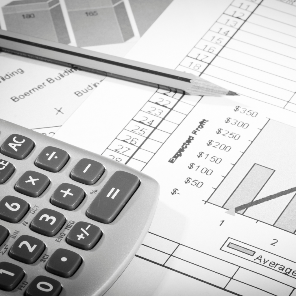 Imagem em preto e branco de uma mesa de trabalho contábil, mostrando uma calculadora em primeiro plano sobre planilhas de dados. Um lápis e gráficos financeiros, incluindo colunas e números de lucro esperado, estão espalhados, simbolizando o cálculo de tributos como a CSLL.