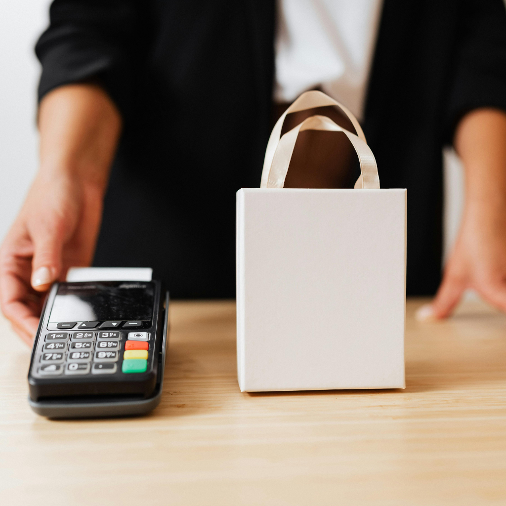 Vendedora seguranda a máquina de passar cartão e ao lado uma sacola da loja. Representando o pagamento da compra.